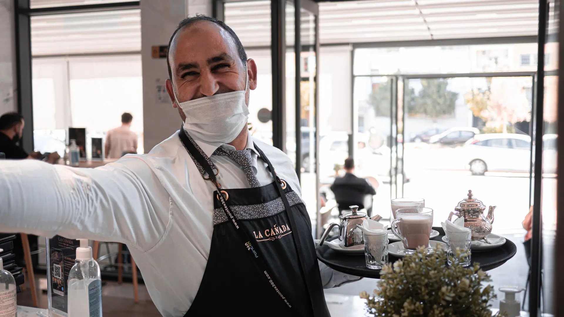 A cheerful waiter with a face mask serves drinks on a tray in a cafe in Fes, Morocco.