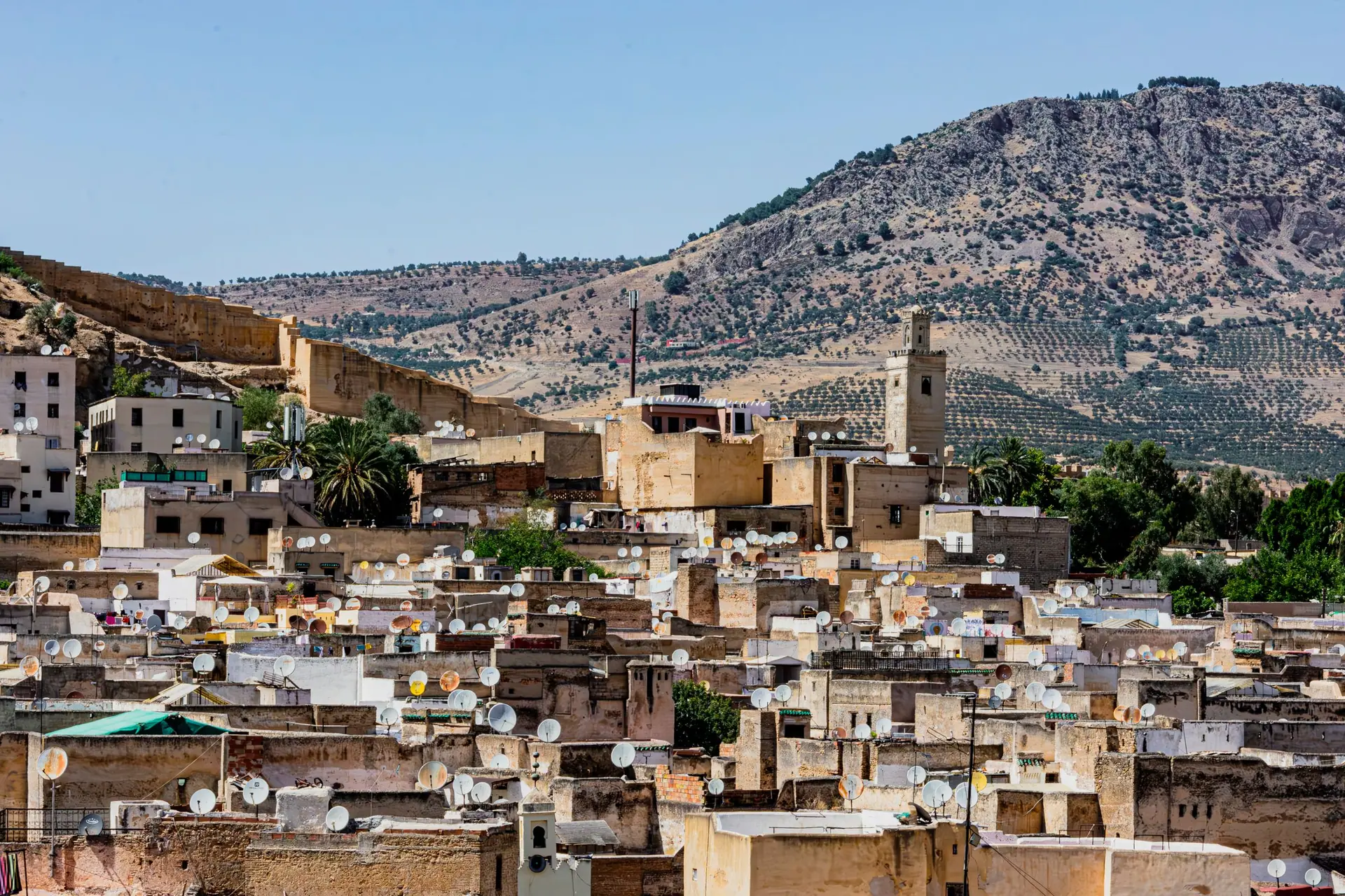 Explore the vibrant rooftops of Fes with satellite dishes, against a backdrop of Moroccan mountains.