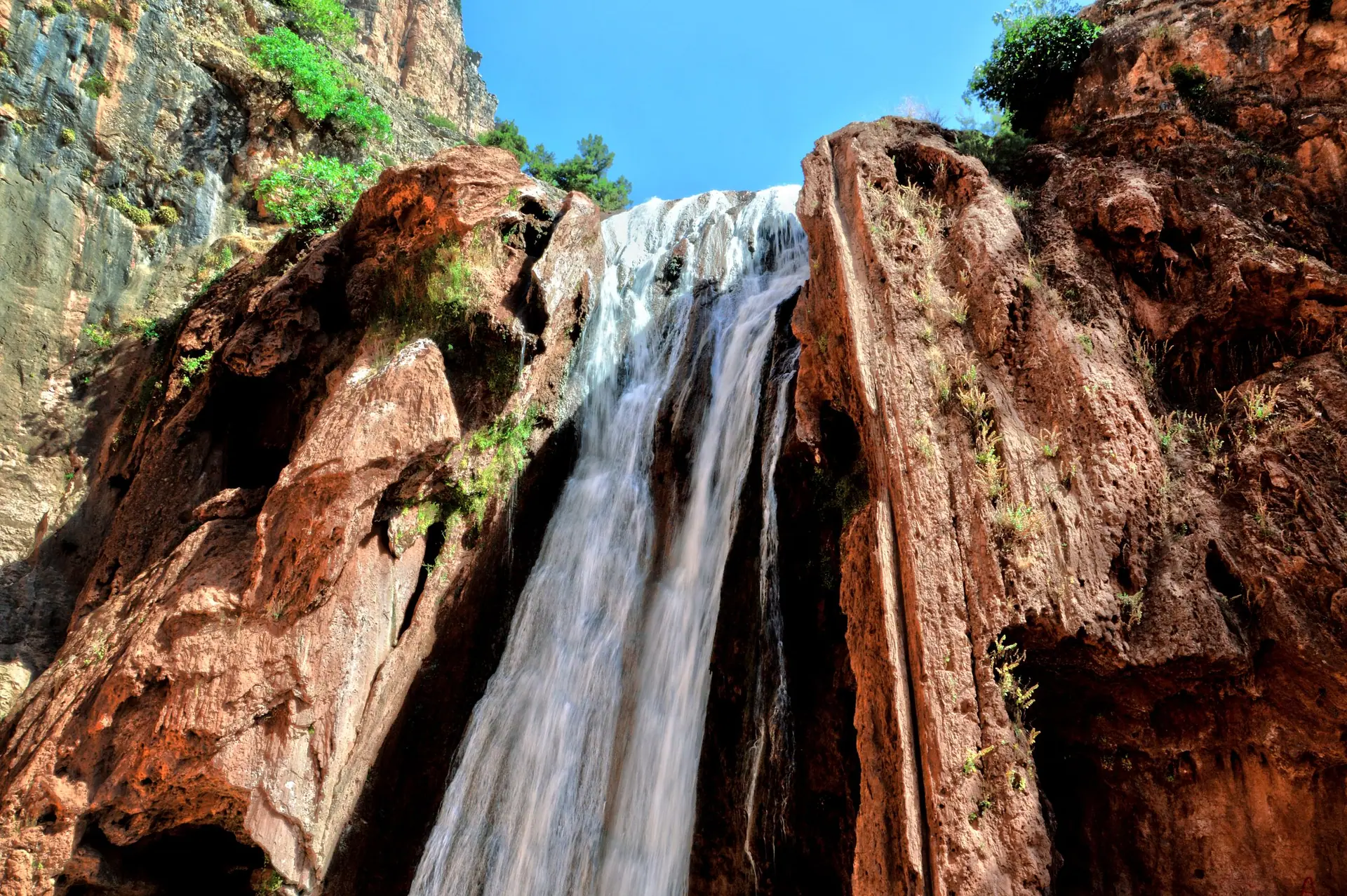 Stunning view of a waterfall cascading down rugged cliffs in Morocco's Atlas Mountains under a clear blue sky.
